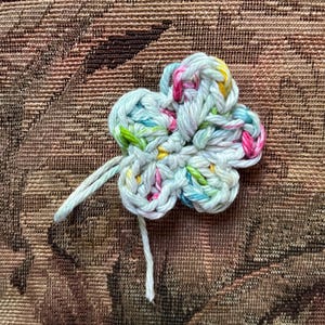 May include: A handmade, crocheted flower in a four-petal design. The flower is made with light blue yarn, with flecks of pink, yellow, and green. Two strands of white yarn extend from the center of the flower.