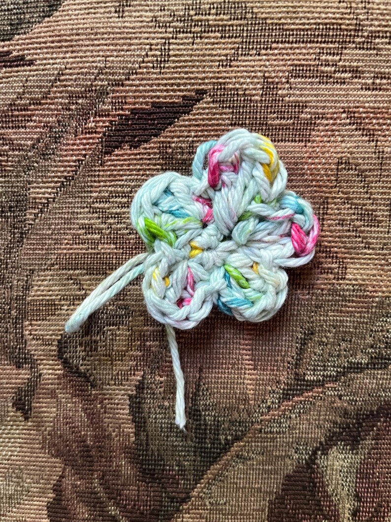 May include: A handmade, crocheted flower in a four-petal design. The flower is made with light blue yarn, with flecks of pink, yellow, and green. Two strands of white yarn extend from the center of the flower.