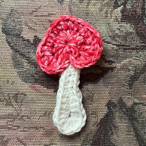 May include: A handmade crochet mushroom with a red and white cap and a white stem. The mushroom is made of yarn and has a textured appearance. The mushroom is a decorative item.