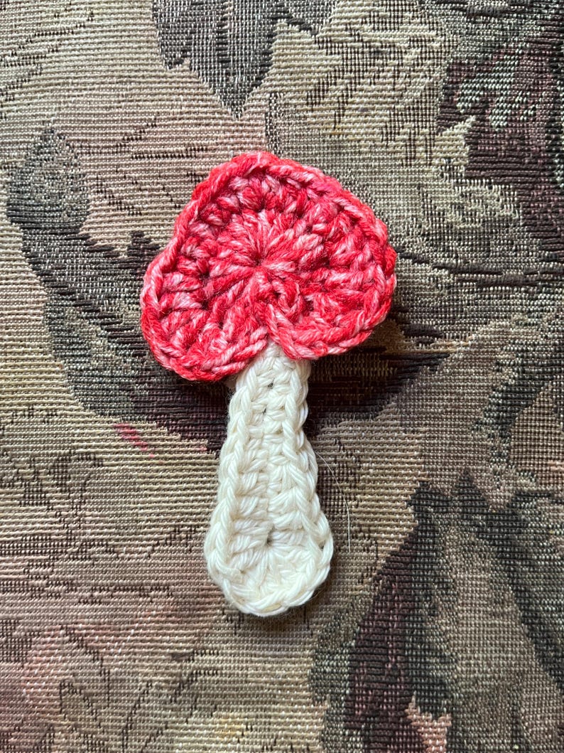May include: A handmade crochet mushroom with a red and white cap and a white stem. The mushroom is made of yarn and has a textured appearance. The mushroom is a decorative item.