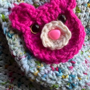 May include: Close-up of a handmade crochet item featuring a bright pink bear face applique. The bear has black eyes, a white flower nose, and a pink heart-shaped nose. The background is a multi-colored crochet fabric.
