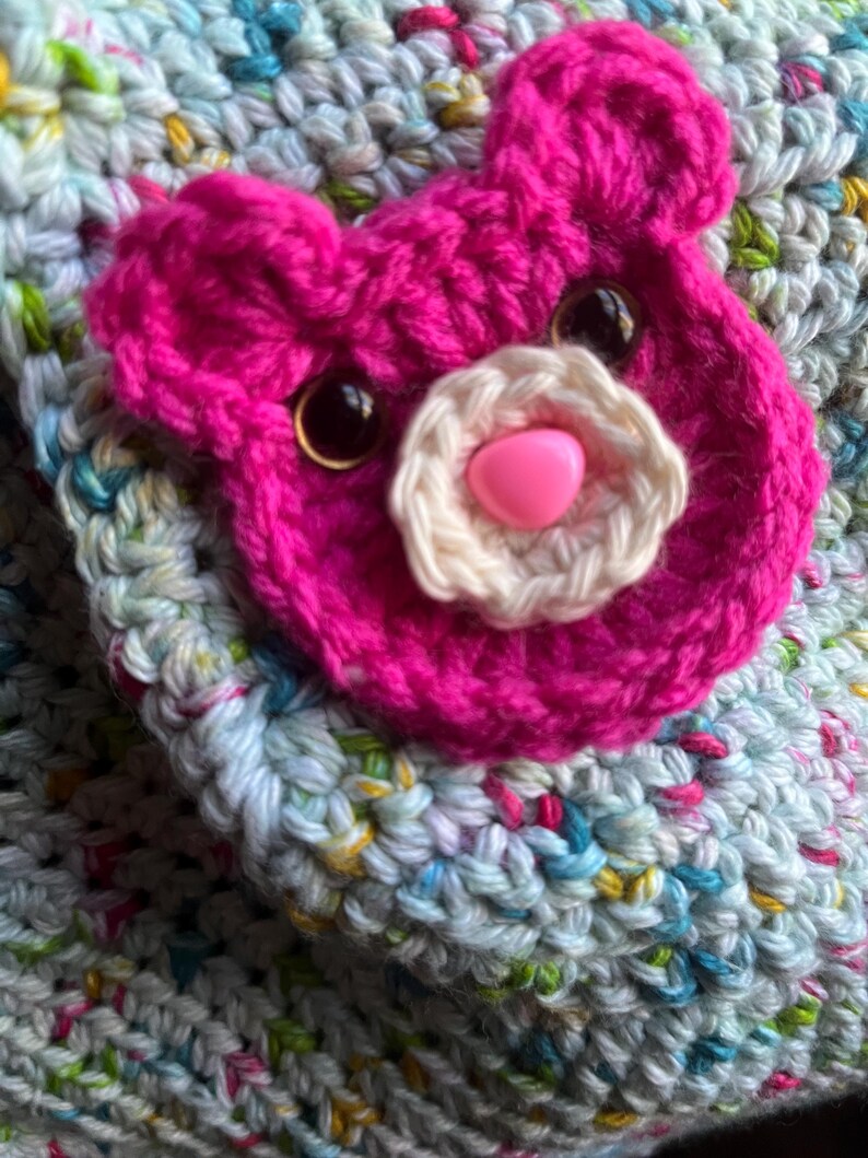 May include: Close-up of a handmade crochet item featuring a bright pink bear face applique. The bear has black eyes, a white flower nose, and a pink heart-shaped nose. The background is a multi-colored crochet fabric.
