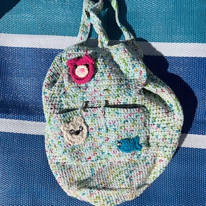 May include: A handmade crochet backpack in a light blue, multi-colored yarn. The backpack features a flap with a pink bear applique, a pocket with a white bear applique, and a blue fish applique. The bag has two straps.