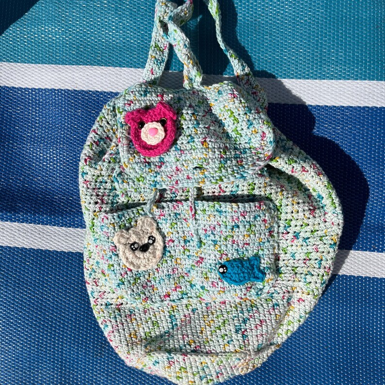 May include: A handmade crochet backpack in a light blue, multi-colored yarn. The backpack features a flap with a pink bear applique, a pocket with a white bear applique, and a blue fish applique. The bag has two straps.