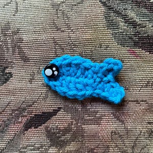 May include: A handmade, crocheted blue fish with a large, black and white button eye. The fish is lying on a patterned, neutral-toned fabric. The fish is a whimsical, handcrafted item.