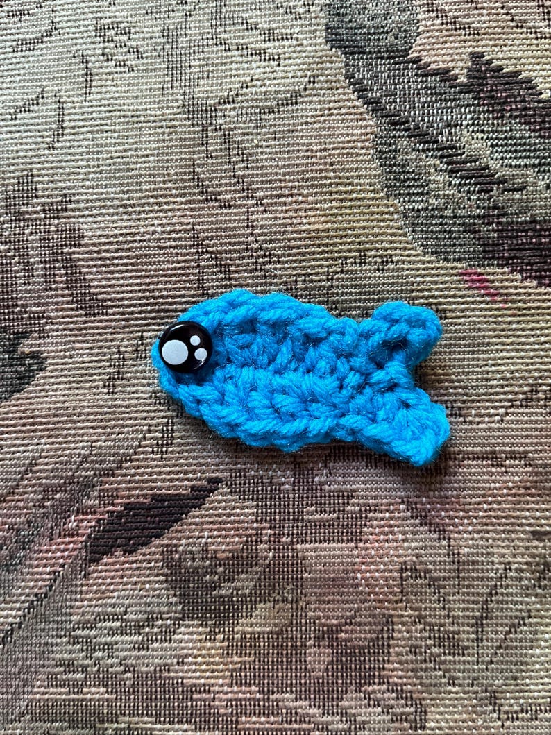 May include: A handmade, crocheted blue fish with a large, black and white button eye. The fish is lying on a patterned, neutral-toned fabric. The fish is a whimsical, handcrafted item.
