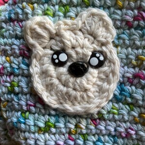 May include: A handmade crochet hat featuring a cream-colored bear face with black and white button eyes and a black nose. The hat is made of multicolored yarn in shades of blue, green, yellow, and pink, creating a textured appearance. The bear face is centered on the hat.