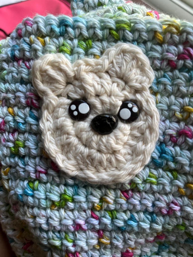 May include: A handmade crochet hat featuring a cream-colored bear face with black and white button eyes and a black nose. The hat is made of multicolored yarn in shades of blue, green, yellow, and pink, creating a textured appearance. The bear face is centered on the hat.
