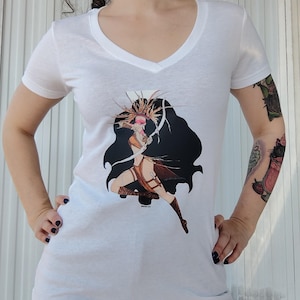 May include: White V-neck t-shirt with a graphic print of a woman with long black hair wearing a headdress and holding a bow and arrow. The graphic is in a silhouette style with a black background.