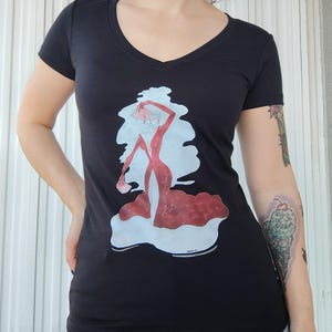 Può includere: Maglietta nera a collo a V con una stampa grafica di una donna in un vestito rosso con una nuvola bianca dietro.