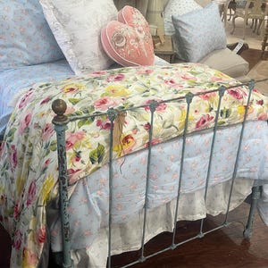 May include: A bed with a floral patterned quilt and light blue sheets. The quilt features pink, yellow, and white roses on a cream background. The bed frame is a distressed blue metal. Decorative pillows in matching floral patterns complete the bedding.