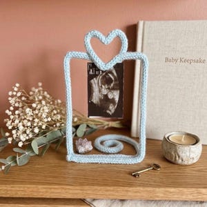 May include: A light blue, knitted picture frame in the shape of a rectangle with a heart at the top, holding a sonogram image. A small candle, a key, and a book titled "Baby Keepsake" are also visible.