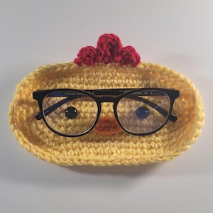 May include: A yellow crocheted eyeglass holder shaped like a chick, with a red comb and an orange beak. Black-framed glasses rest inside the holder. The holder is designed to keep glasses safe and secure.