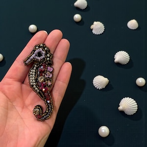 May include: A handcrafted seahorse brooch with intricate beadwork in shades of brown, silver, and red. The seahorse is embellished with sequins and small beads. White seashells and pearls are scattered around the brooch on a dark teal surface.