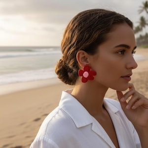 Op de afbeelding: Een vrouw draagt grote, rode bloemvormige oorbellen met een roze centrum. De oorbellen zijn het middelpunt, tegen een strandachtergrond met de oceaan en de lucht zichtbaar. De vrouw draagt een wit overhemd met kraag.