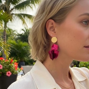 Może przedstawiać: Kolczyki z pozłacanymi, teksturowanymi dyskami i warstwowymi, płatkowatymi zawieszkami w głębokim odcieniu magenty. Kolczyki stanowią wyrazisty element, dodający koloru i faktury.
