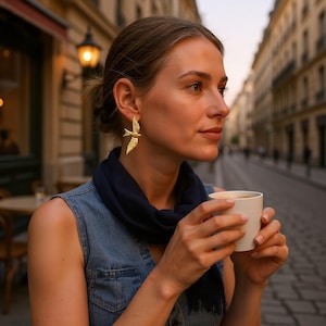 May include: A woman wearing gold bird-shaped earrings. The earrings are detailed with textured wings and a delicate design. She is wearing a denim vest and a dark blue scarf, holding a white mug in an outdoor setting.