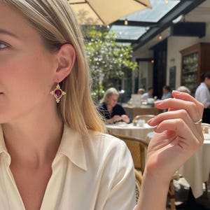 May include: Gold-toned drop earrings with a teardrop-shaped, deep red gemstone center. The earrings feature a cross-like design with small, round accents. The setting is a warm gold color, and the earrings are worn on a person.