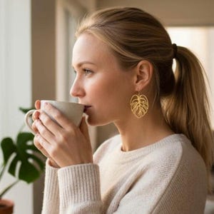 Op de afbeelding: Een vrouw draagt gouden oorbellen in de vorm van een blad en een crèmekleurige trui. Ze drinkt uit een witte mok. Haar blonde haar is in een paardenstaart gebonden. Op de achtergrond is een groene plant te zien.
