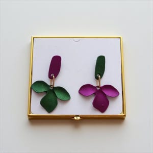 May include: A pair of floral-shaped earrings. One earring features a green petal design with a dark purple top, while the other has a purple petal design with a green top. Both earrings are attached with gold-colored hoops.