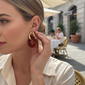 May include: A drop earring featuring two interlocking oval links. The top link is crafted from polished gold-tone metal, while the bottom link is made of brown tortoiseshell-patterned resin. The earring hangs vertically from the earlobe.