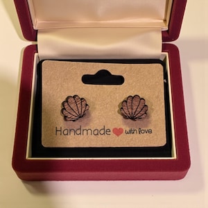 Puede incluir: Pendientes de botón de concha de madera presentados en una caja de joyería burdeos. Los pendientes son marrones con detalles negros. Están sobre una tarjeta marrón que dice "Handmade with love".