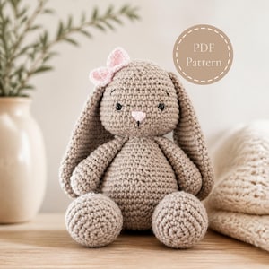 Può includere: Un coniglietto di peluche all'uncinetto di colore marrone chiaro con orecchie lunghe, un piccolo naso rosa e un fiocco rosa chiaro attaccato al lato della testa. Il giocattolo presenta occhi ricamati neri e un badge circolare con il testo PDF Pattern.