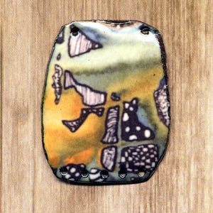 May include: A colorful, abstract design on a copper-toned metal pendant with a textured surface. The pendant has a slightly irregular shape and is decorated with a variety of colors, including orange, yellow, green, black, and white.