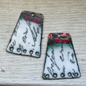 May include: Two trapezoidal metal pendants featuring a white enamel surface with black cursive script text. The top edges are finished in a dark, textured copper-toned patina with red accents, and each piece includes five small circular holes.