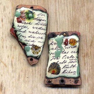 May include: Two handmade, irregular-shaped ceramic pendants. Each pendant features a white background with handwritten text, colorful floral and abstract designs, and a hole for hanging. The edges are brown with red accents.