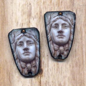 May include: Two pendant-style charms with a classical female face design. The pendants are rectangular with a rounded bottom and a hole at the top. The faces are in relief, with detailed hair and a bow at the neck, in shades of white, gray, and brown.