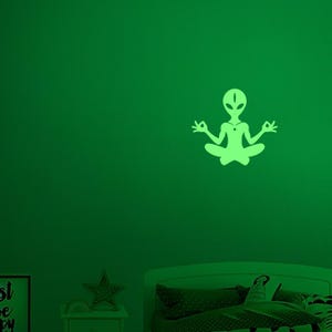 May include: A glowing green alien silhouette in a seated yoga pose, with hands in a mudra. The alien has a large head and a necklace. The image is on a green wall above a bed and nightstand.