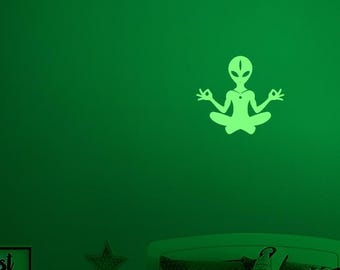May include: A glowing green alien silhouette in a seated yoga pose, with hands in a mudra. The alien has a large head and a necklace. The image is on a green wall above a bed and nightstand.