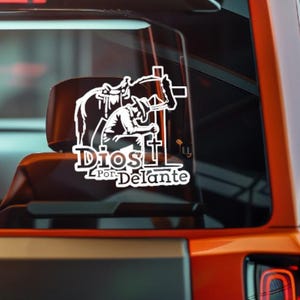 May include: White decal featuring a cowboy kneeling before a cross, with a horse and saddle in the background. The text "Dios Por Delante" is below the image. The decal is likely for a vehicle window.