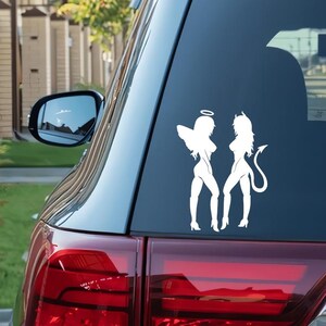 May include: White vinyl decal featuring an angel and a devil silhouette. The angel has wings and a halo, while the devil has horns and a tail. The decal is applied to the rear window of a vehicle.