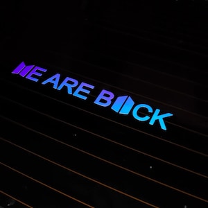 May include: A car window decal with the words "WE ARE BACK" in a gradient of blue and purple. The text is applied to the rear window of a vehicle.