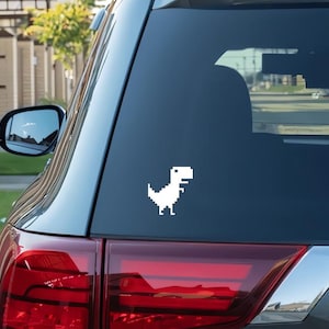 May include: A white pixelated dinosaur decal is affixed to the rear window of a vehicle. The dinosaur is in a side-facing pose, with its head and tail raised. The decal is a simple, retro design.