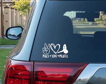 May include: White vinyl decal applied to a car window. The decal features a peace sign, a heart, and a pilates pose, with the words "PEACE LOVE PILATES" below. The car is silver.