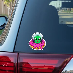 May include: A green alien with a pink donut spaceship. The donut has sprinkles and pink icing.