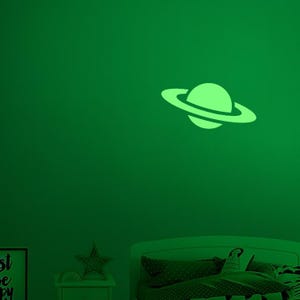 May include: A glowing green Saturn-shaped wall decal. The planet has a circular ring around it, and is affixed to a green wall. The decal is part of a space-themed room decor.