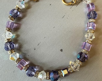 May include: A purple, white, and clear crystal beaded bracelet with a gold clasp. The bracelet features a variety of crystal shapes and sizes.