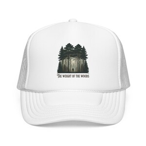 May include: A white trucker hat featuring a graphic print of a dark forest with a small silhouette of a person standing among the trees. Below the forest illustration, the text reads THE WEIGHT OF THE WOODS in a dark, stylized serif font.