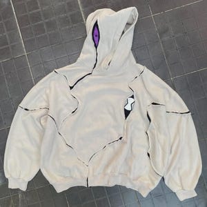 May include: Cream-colored hoodie with a unique design featuring black stitching and a hood. The hood has a purple eye detail, and the front has a black and white graphic. The hoodie has a relaxed fit and long sleeves.