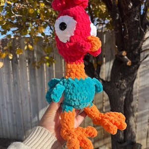 May include: A crocheted plush toy rooster with a red head, teal body, and orange legs. The rooster has large white eyes with black pupils.