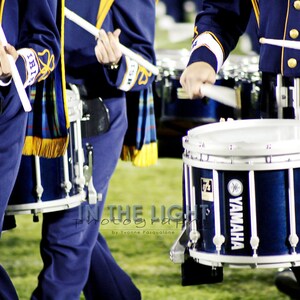 May include: A marching band drum with a blue and white finish. The drum has the word "YAMAHA" printed on the side. The drum is being held by a person wearing a blue uniform.