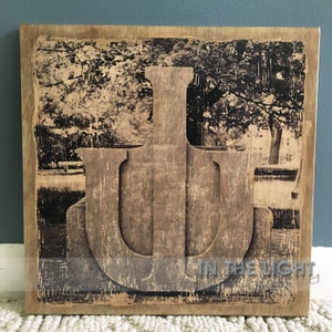 May include: A wooden sign with a distressed finish featuring a large, carved letter "U" in the center. The background is a faded black and white photograph of trees and a bench. The text "IN THE LIGHT" is printed in the bottom right corner.