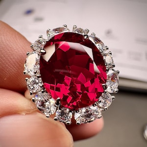 May include: A silver ring featuring a large, oval-cut, vibrant red gemstone as the centerpiece. The central stone is surrounded by a halo of smaller, clear, round-cut gemstones, creating a sparkling, elegant design. The ring is set against a neutral background.