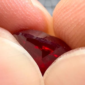 May include: Close-up of a deep red gemstone with a faceted cut. The gemstone is held between two fingers and the word "remarkable" is inscribed on the surface.