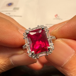 May include: A silver-toned ring featuring a large, rectangular, deep red centre stone. The centre stone is surrounded by a halo of smaller, clear, baguette-cut and round-cut stones. The band is thin and accented with small, clear, sparkling stones.