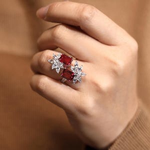 May include: A silver ring with two red gemstones and white diamond accents. The ring features a floral design.